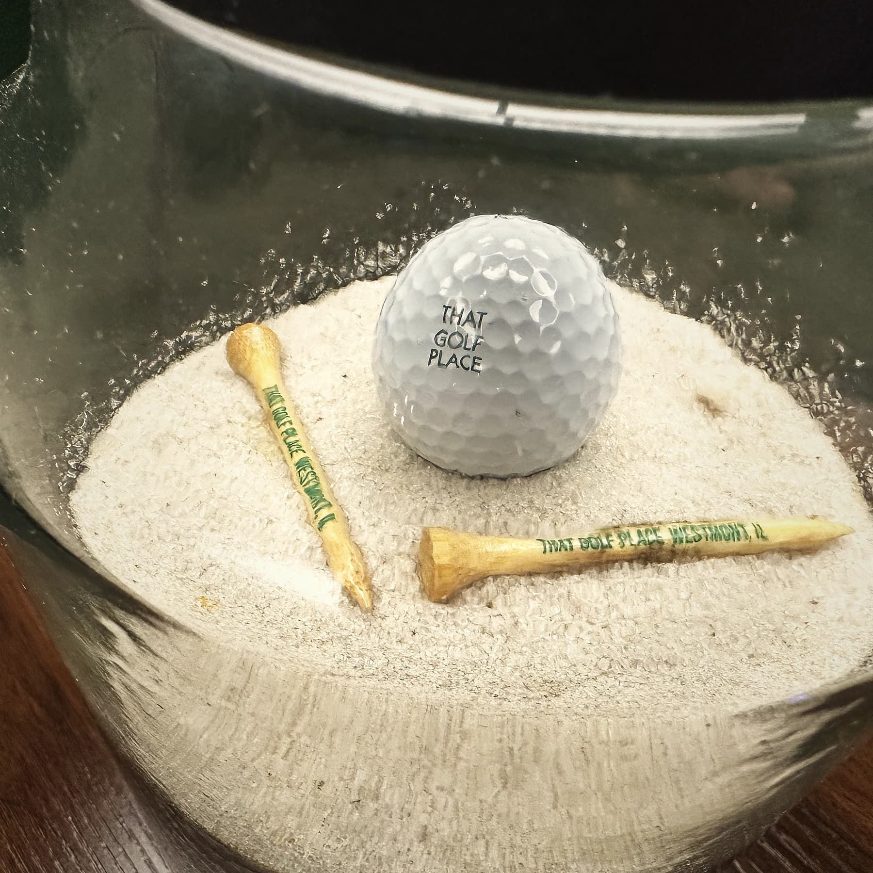 Branded That Golf Place golf ball resting in a sand-filled tee jar with two branded wooden tees stamped 'That Golf Place Westmont, IL'