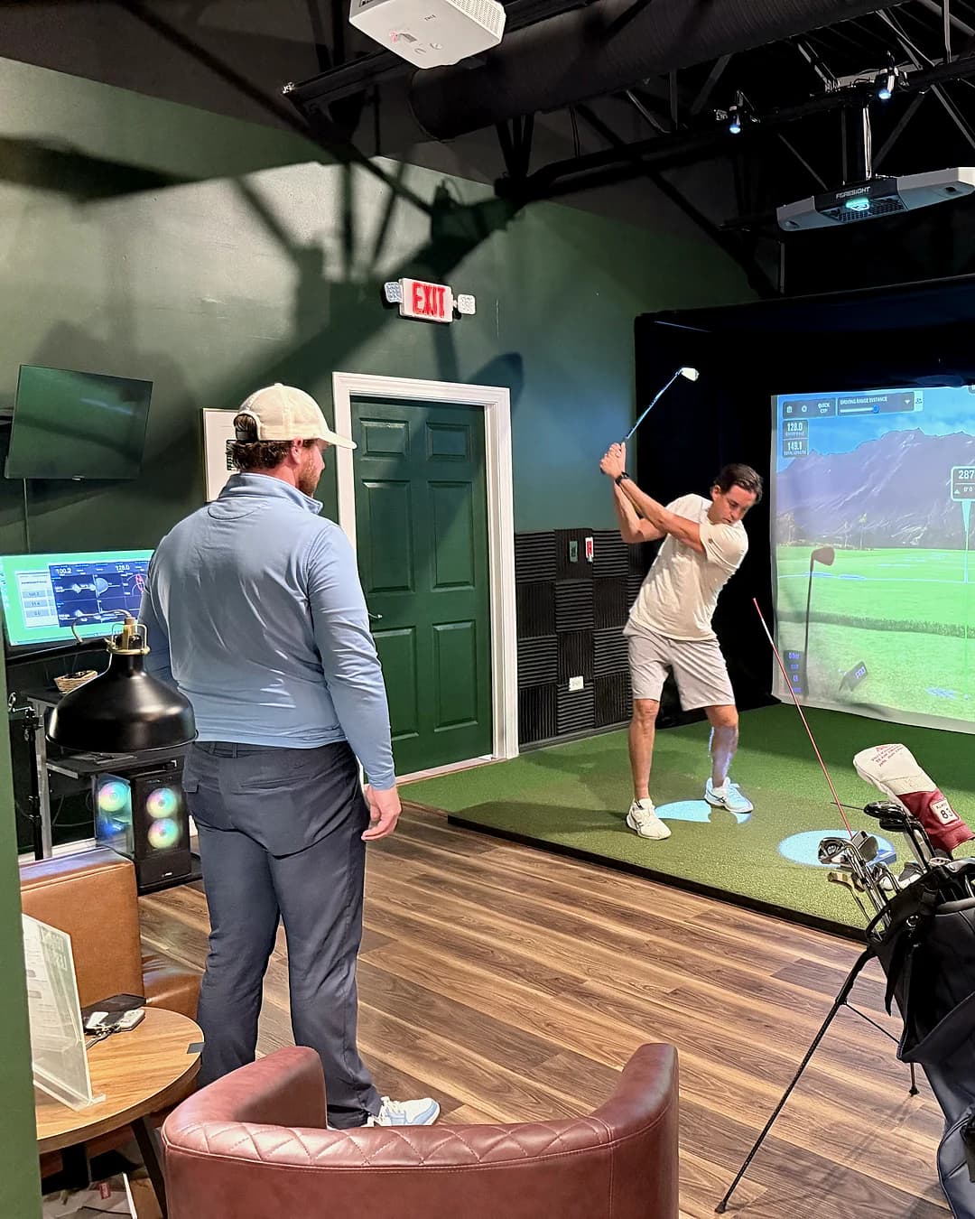 A coach observing a player mid-backswing inside a private simulator bay at That Golf Place, both figures silhouetted against the lit GSPro screen