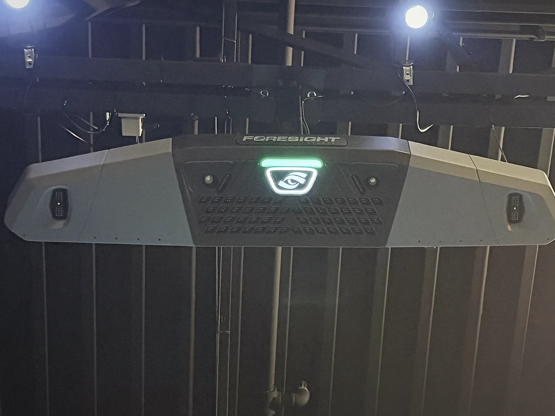 Foresight Falcon launch monitor mounted overhead between two stage lights in a bay at That Golf Place, glowing green logo against deep moody shadows